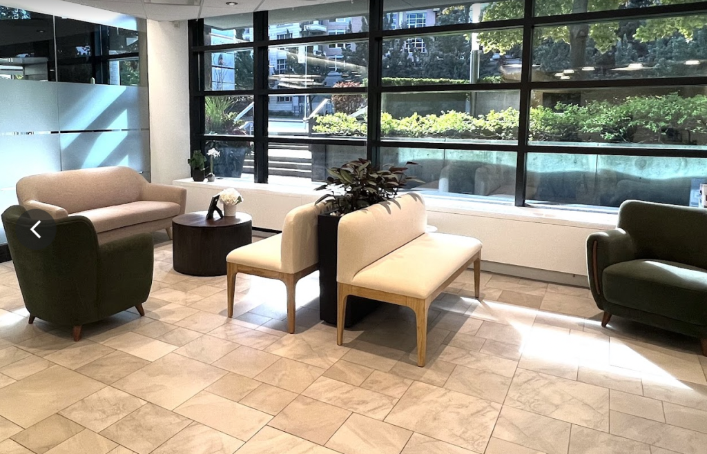 Medical waiting room with nice beige chairs, light coloured floor, large window open to the street with lots of greenery